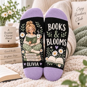 May include: A pair of black socks with lavender cuffs. The left sock features an illustration of a person reading a book while holding a mug, sitting on a stack of books labelled Good Books, Brighter Days, Happier Me, and the name OLIVIA. The right sock displays the text BOOKS & BLOOMS with floral accents and an open book.