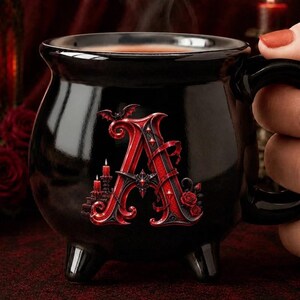 Puede incluir: Una taza de cerámica negra con forma de caldero que presenta una letra A roja grande y ornamentada en el frente. La letra está decorada con pequeños murciélagos, velas encendidas y rosas rojas. La taza tiene tres patas cortas y un asa.