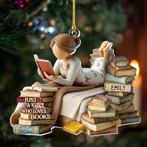 May include: A decorative ornament of a person reading a book while lying on a bed of books. The ornament has the text "JUST A GIRL WHO LOVES BOOKS" and the name "EMILY" on the books. The person is wearing a white floral patterned outfit.
