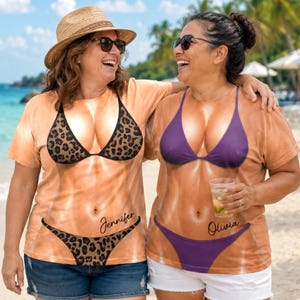 Może przedstawiać: Dwie koszulki z krótkim rękawem i okrągłym dekoltem z realistycznym nadrukiem tułowia w kolorze skóry w bikini. Jedna koszulka przedstawia bikini w panterkę z imieniem Jennifer, a druga gładkie fioletowe bikini z imieniem Olivia.