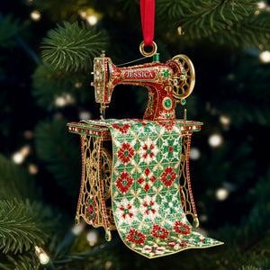 May include: A red and gold sewing machine Christmas ornament with the name "Jessica" on top. The ornament features intricate details, including a patterned fabric draped over the table. The ornament is hanging from a red ribbon against a blurred Christmas tree background.