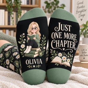 May include: A pair of black socks with sage green cuffs. One sock features a cartoon character with glasses reading a book, surrounded by daisies and the name OLIVIA. The other sock displays the text JUST ONE MORE CHAPTER above an open book.
