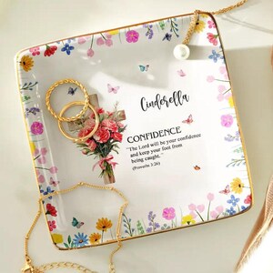 以下が含まれることがあります： 金色の縁取りと花のボーダー、そして「Cinderella」と「CONFIDENCE」の文字が入った正方形の白いセラミック製の小物入れ。お皿には、花、蝶、金の指輪が付いた十字架が描かれています。テキストには「主はあなたの自信となり、あなたの足を捕らわれることから守ってくださる。」と書かれています。（箴言3:26）。
