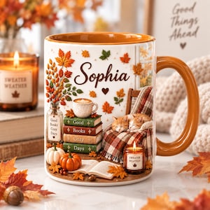 May include: A white ceramic mug with an orange interior and handle, featuring the name Sophia in script, a stack of books labeled Good Books Cozy Days, two sleeping orange cats on a plaid blanket, a coffee cup, pumpkins, and autumn leaves.