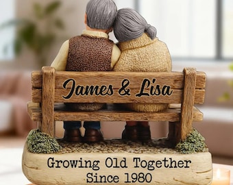 May include: A figurine of an elderly couple sitting on a wooden bench, with the names "James & Lisa" carved into the backrest. The base reads "Growing Old Together Since 1980". The couple is wearing sweaters and vests.