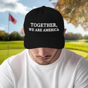 May include: Black baseball cap with the text "TOGETHER, WE ARE AMERICA" in white. The cap is worn by a person in a white shirt, with a golf course in the background.