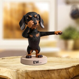 May include: A Dachshund figurine in a disgusted pose, holding its nose with one paw and extending the other. The dog is black with tan markings and stands on a beige base with the text "EW...".
