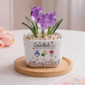 Op de afbeelding: Een witte keramische pot met drie paarse glazen tulpenbloemen. De pot heeft de tekst "My Grandkids make my life more grand" en is versierd met bloemenillustraties en namen. Hij staat op een ronde houten schaal.