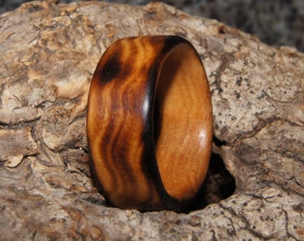 Puede incluir: Un anillo de madera con un patrón de veta de madera marrón oscuro y marrón claro. El anillo está hecho de madera natural y tiene un acabado liso y pulido.