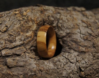 Puede incluir: Un anillo de madera con un patrón de rayas marrón claro y beige. El anillo está colocado sobre un fondo de madera marrón texturizado. El anillo parece estar hecho a mano y tiene un acabado liso y pulido.
