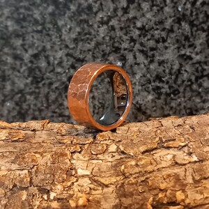 Puede incluir: Un anillo de color cobre con una textura martillada. El anillo es ancho y tiene un interior oscuro. El anillo se muestra sobre un trozo de madera contra un fondo oscuro. El anillo es una joya.