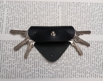 May include: A black leather key holder with a snap closure and a silver button. The holder is holding a set of keys with silver metal.