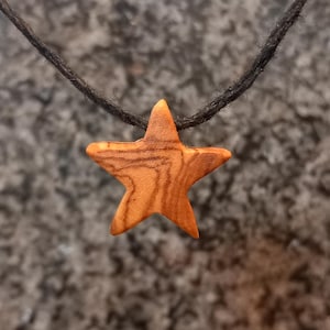 Può includere: Una collana con pendente a forma di stella in legno. La stella è di colore marrone caldo con un motivo a venature del legno visibile. Il cordino della collana è un materiale scuro e intrecciato. Il pendente è sospeso su uno sfondo sfocato e strutturato.
