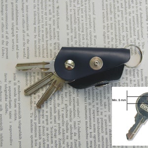 May include: A close-up of a silver key with a black leather key holder. The key has a black plastic head with the text "SKS" printed on it. The key holder has a metal ring and two metal rivets. The key is 5 mm wide.