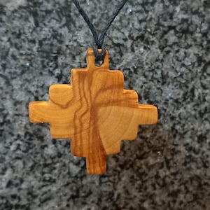 May include: A light brown wooden pendant shaped like a stepped cross with a visible natural grain pattern. The pendant is suspended from a thin black cord that passes through a small hole at the top center of the geometric wooden structure.