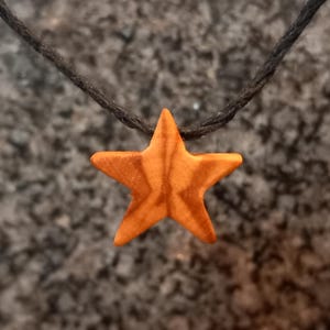Può includere: Una collana con pendente a forma di stella in legno. La stella è di colore arancione caldo ed è sospesa a un cordino nero. La stella ha una superficie strutturata e ha una forma a cinque punte. La collana è un design semplice e minimalista.