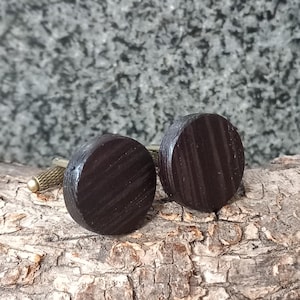 May include: A pair of dark brown wooden cufflinks with a circular design. The cufflinks have a textured, striped pattern and are set on a gold-colored metal base. The background is a textured, gray surface.
