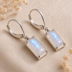 May include: A pair of silver-toned leverback earrings featuring rectangular, faceted moonstone gems. Each stone displays a translucent white appearance with subtle blue iridescent flashes, securely held in a four-prong metal setting.