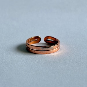 May include: A copper-toned metal ring featuring a double-band design with an open gap at the top. The ring has a smooth, polished finish and a simple, continuous structure that wraps around to form a circular shape with a visible opening.