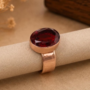 May include: A ring featuring a large, oval-cut, deep red gemstone set in a polished rose gold band. The faceted stone is securely held within a smooth metal bezel setting, showcasing a vibrant crimson hue and a reflective, high-shine finish.