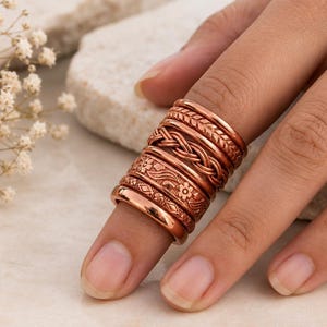 May include: A stack of five copper-toned metal rings worn on a finger. The rings feature various textures and patterns, including a smooth band, a geometric diamond design, a floral motif, a braided rope texture, and a diagonal etched pattern.