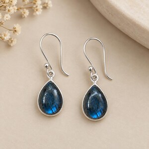 Puede incluir: Un par de pendientes colgantes de plata con piedras de labradorita en forma de lágrima. Cada pendiente tiene un gancho de plata pulida y un engaste que sujeta la piedra preciosa azul oscuro e iridiscente con destellos naturales.