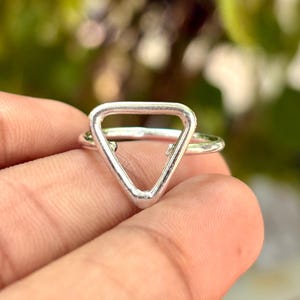 Peut inclure: Une bague de couleur argentée présentant une forme triangulaire ouverte proéminente comme point focal. L'anneau est fin et circulaire, se connectant à la base du triangle. Le métal a une finition lisse et polie qui reflète la lumière.