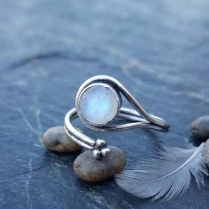 May include: A silver ring featuring a round, translucent moonstone cabochon set in a polished metal frame. The band has an open, bypass-style design that tapers into a decorative cluster of three small metal spheres at the terminal end.