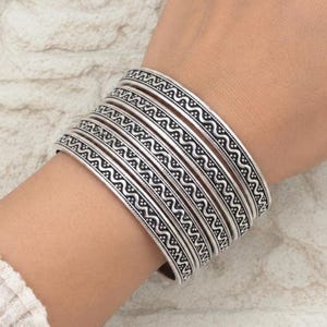 Può includere: Un set di cinque bracciali rigidi in metallo color argento indossati insieme. Ogni bracciale presenta un motivo costante con una linea ondulata centrale affiancata da file di piccoli punti, con dettagli neri ossidati nei solchi incisi.