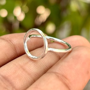 Puede incluir: Un anillo de plata con un diseño de marco abierto circular. La banda es fina y lisa, conectada a un aro de metal pulido y redondo que se asienta verticalmente. El metal tiene un acabado brillante y reflectante, con una estética simple y geométrica.