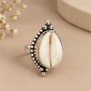May include: A silver ring featuring a central white cowrie shell set within a teardrop-shaped frame. The metal border is decorated with a series of small, raised silver spheres, creating a textured, beaded effect around the shell.