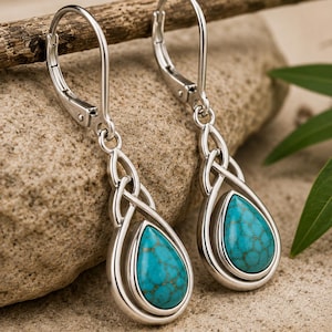 May include: A pair of silver-toned drop earrings featuring a teardrop-shaped turquoise stone with brown veining, set within a polished metal frame. The design includes an intricate interwoven metal knot detail above the stone and a lever-back closure.