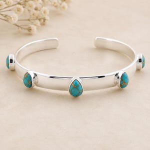 May include: A silver cuff bracelet featuring five teardrop-shaped turquoise stones with brown matrix patterns. The polished metal band is open at the back, and the stones are evenly spaced along the front and sides of the smooth, reflective surface.