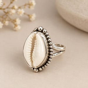 May include: A silver ring featuring a central white cowrie shell set within a beaded metal border. The band is constructed from two smooth silver wires that merge into a single shank, providing a polished finish to the overall jewellery piece.