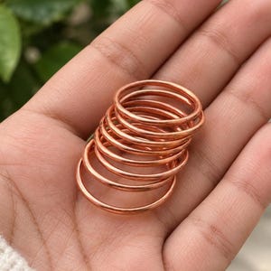 May include: A stack of seven thin, smooth, circular copper rings with a polished metallic finish. The rings are uniform in thickness and color, featuring a simple, unadorned design that highlights the warm, reddish-brown tone of the metal.