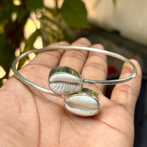 Puede incluir: Una pulsera rígida de tono plateado que presenta dos conchas de cauri blancas engastadas en biseles de metal en los extremos abiertos. La pulsera tiene un acabado liso y pulido, con las conchas posicionadas una frente a la otra en la abertura.