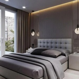 May include: A modern bedroom with a gray upholstered bed, featuring a tufted headboard and layered bedding. The room has a neutral color palette, with gray walls, curtains, and a wooden floor. Decorative pendant lights hang from the ceiling.
