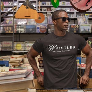 May include: A black t-shirt with the white text "Whistles Pub & Eatery - Scranton Pa." The shirt is worn by a person wearing sunglasses, standing in a shop filled with books and records.