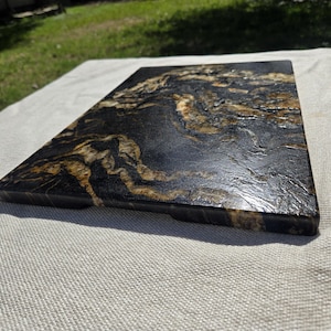 May include: A rectangular serving board featuring a high-gloss finish with a marbled pattern of deep black and metallic gold swirls. The surface has a textured appearance with visible depth and organic, flowing lines across the entire top face.