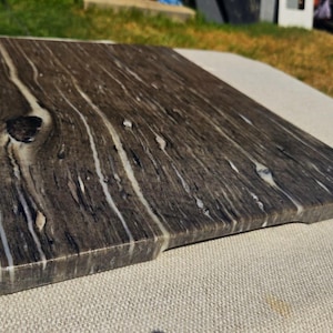 May include: A rectangular serving board made of dark gray stone with prominent white veining and a distinct dark knot feature. The surface has a polished finish, showcasing natural linear patterns that run across the length of the board.