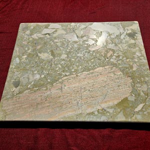 May include: A square slab of polished stone featuring a variegated pattern of sage green, olive, and cream-colored mineral fragments. A large, elongated section of light pink and tan veined stone is embedded diagonally across the lower right surface.