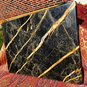 Puede incluir: Una losa cuadrada de mármol negro pulido con llamativas vetas doradas. La piedra natural presenta un acabado brillante y un patrón único de vetas doradas en su superficie. La losa probablemente se utiliza con fines decorativos o como encimera.