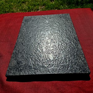 May include: A rectangular slab of dark gray granite with a rough, textured surface. The stone features natural mineral flecks and a polished finish that reflects light across its uneven, craggy top surface, showcasing a solid, heavy appearance.