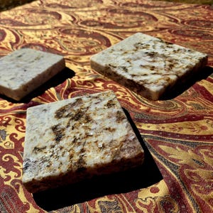 May include: Three square stone coasters with a speckled white and brown pattern. The coasters are arranged on a red, gold, and brown paisley patterned fabric. The coasters are approximately 4 inches square.