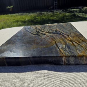 May include: A rectangular stone serving board featuring a polished surface with a natural marbled pattern in shades of deep charcoal, slate gray, and warm golden brown, showcasing intricate veining throughout the entire flat top.
