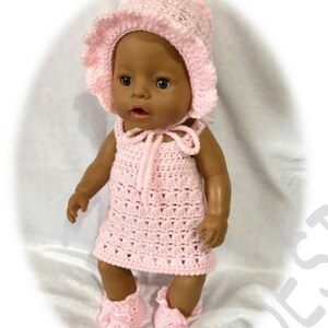Könnte beinhalten: Ein pinkfarbenes Häkel-Outfit für eine Baby-Puppe, bestehend aus einem Kleid, einer Mütze und Schühchen.