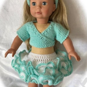 Op de afbeelding: Pop gekleed in een handgemaakt gehaakt outfit. De outfit bestaat uit een turquoise wikkel top, een wit en turquoise gerokte rok en een bijpassende hoofdband. De pop draagt ook witte sneakers.