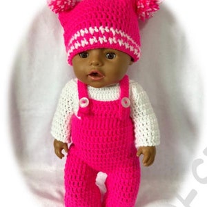 Puede incluir: Un conjunto de muñeca de crochet rosa con un gorro a juego. El gorro tiene pompones blancos y rayas blancas. El conjunto incluye una camisa blanca de manga larga y un mono rosa.