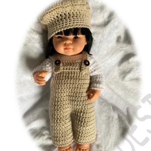 May include: A crocheted doll outfit in beige with a matching beret. The outfit includes a long-sleeved shirt and overalls. The doll has black hair and brown eyes.