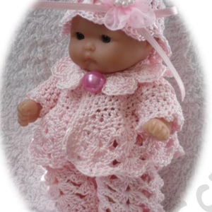 May include: A pink crocheted outfit for a doll, including a hat, sweater, and pants. The outfit features delicate crocheted details and a white flower embellishment on the hat.